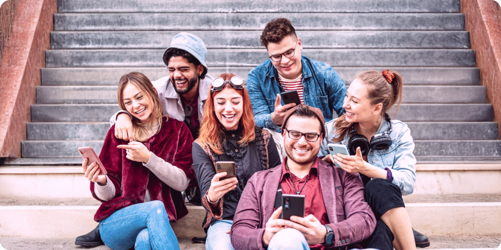 Friends enjoying time together while using mobile phones "A group of six young adults sitting on outdoor stairs, smiling and laughing while looking at their smartphones together."
